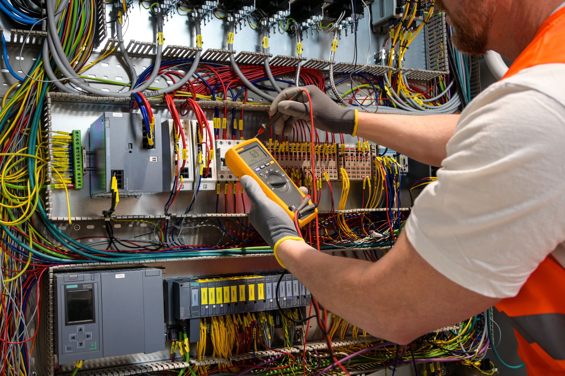 an electrician in a cap, yellow glasses and an orange vest measures electric current with a digital multimeter on a distribution box